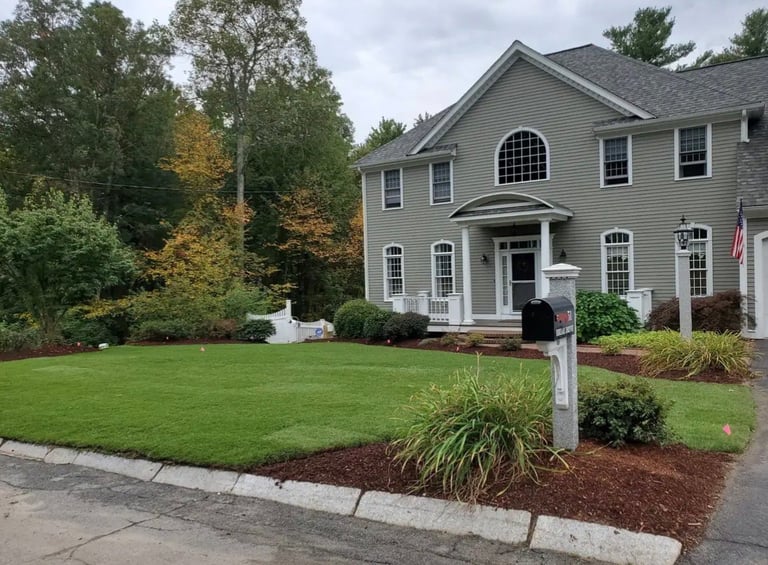 Two-story gray colonial house with manicured lawn, landscaped garden beds, and trees in the background on a residential street