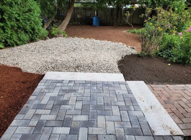 Backyard landscaping with gray paver patio, white gravel, dark mulch, and surrounding garden beds with plants