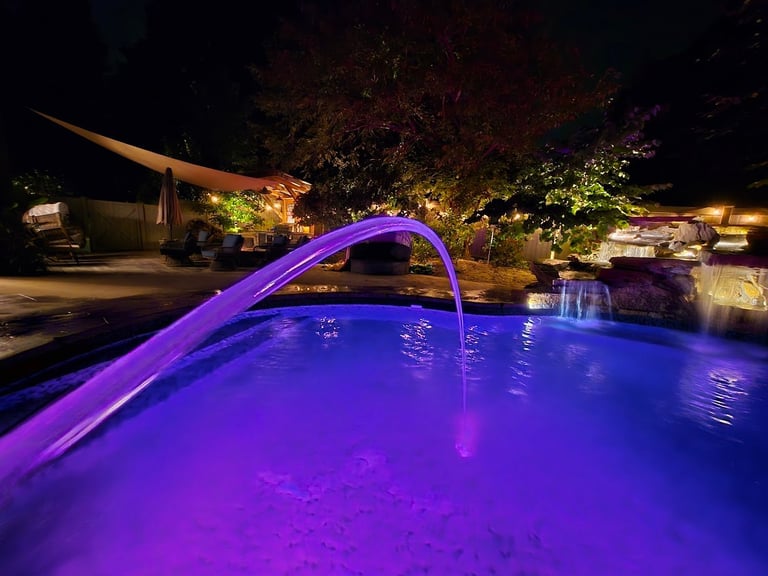 Swimming pool with vibrant purple LED lighting at night, featuring an arched water fountain and illuminated surrounding landscape