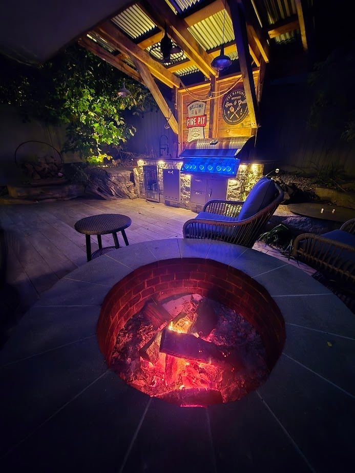 Nighttime backyard with glowing red fire pit, wooden pergola with warm lighting, lounge chairs, and decorative signage