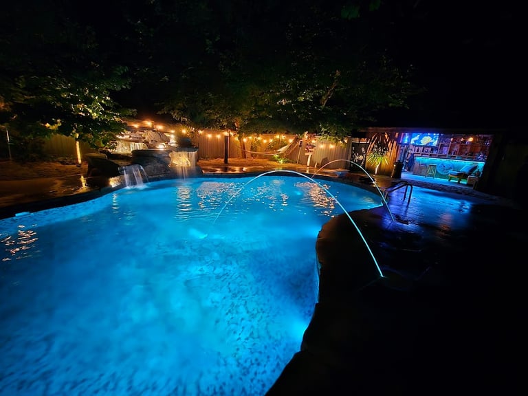Illuminated backyard pool at night with glowing blue water, surrounded by warm landscape lighting and trees