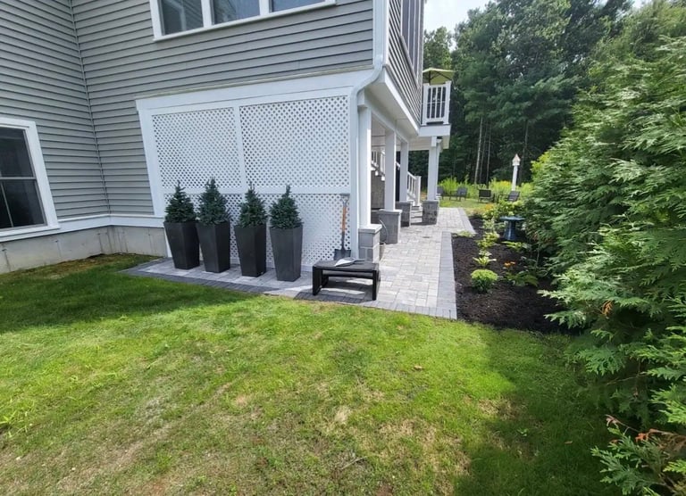 Modern house with white lattice screen, potted evergreen topiaries, paved patio area, and manicured green lawn with shrubs