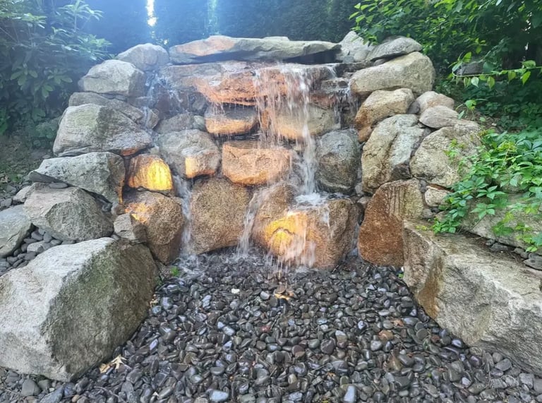 Stone waterfall feature with stacked rocks and water cascading down, surrounded by green foliage and gravel base